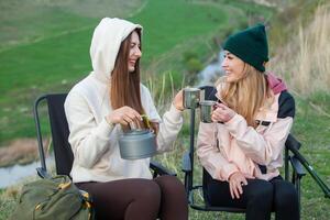 två skön flickor Sammanträde i en hopfällbar stol och dricka te i natur. resa, resande. foto