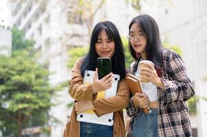 två asiatisk kvinna högskola studenter är stående tillsammans i de campus parkera, ser på en smartphone. foto