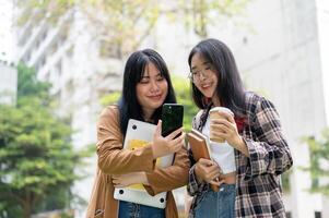 två asiatisk kvinna högskola studenter är stående tillsammans i de campus parkera, ser på en smartphone. foto