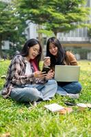 två högskola studenter är Sammanträde på gräs i en parkera, diskuterar något på en smartphone tillsammans. foto