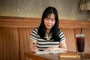 en ung asiatisk kvinna är skrivning henne dagbok eller tar anteckningar i henne anteckningsbok på en kaffe affär. foto