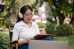 en ung asiatisk kvinna är fokusera på skrivning henne arbete eller förvaring en dagbok medan Sammanträde i en bakgård. foto