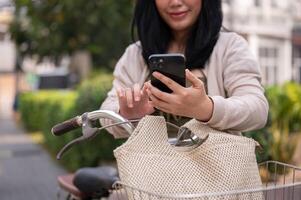 en skön, Lycklig ung asiatisk kvinna i en söt klänning är använder sig av henne smartphone på henne cykel i de stad foto