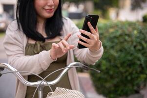 en skön, Lycklig ung asiatisk kvinna i en söt klänning är använder sig av henne smartphone på henne cykel i de stad foto