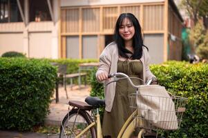 en leende ung asiatisk kvinna i en söt klänning med henne cykel i de stad på en solig dag. foto