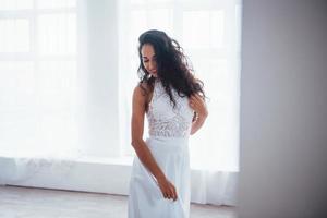 underbart porträtt. vacker kvinna i vit klänning står i vitt rum med dagsljus genom fönstren foto