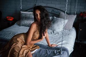 vackert svart hår. flicka i gyllene klänning sitter vid vit säng på kedjorna i lyxiga lägenheter