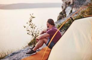 turist i shorts och skjorta sitter vid kanten av berget framför tältet foto