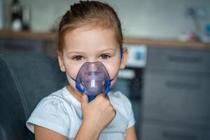 söt liten flicka är Sammanträde och innehav en nebulisator mask lutande mot de ansikte, luftväg behandling begrepp foto