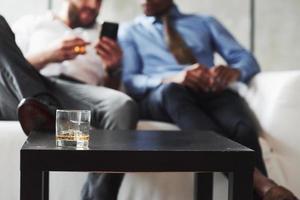 fokusera på det nästan tomma whiskyglaset med is. internationella kollegor som sitter i den vita soffan och tittar på affärsgrejer foto