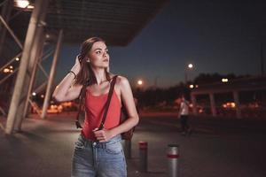 en ung vacker flicka med en ryggsäck bakom axlarna står på gatan nära en flygplats eller en järnvägsstation en varm sommarkväll. hon har precis kommit och väntar på en taxi eller sina vänner foto
