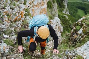 en turist klättrar in i de bergen klängande till stenar med hans händer, en tung ryggsäck av en turist på hans tillbaka, en gul värma hatt, klättrande, en man i de berg. foto