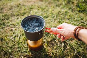 hand innehar en pott av varm vatten, en turist kockar mat på en vandra på en gas brännare, turist redskap, camping i de skog, koka upp vatten för te. foto
