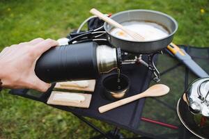 hand innehav termos häller te in i en mugg, turist kök i camping i natur, fräsning panorera med krypterade ägg, mat på en vandra, bergsklättring avslappning foto