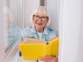 äldre kvinna med grått hår läser en bok vid fönstret hemma. utbildning, pension, anti ålder, läsning koncept