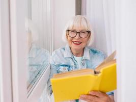 äldre kvinna med grått hår läser en bok vid fönstret hemma. utbildning, pension, anti ålder, läsning koncept