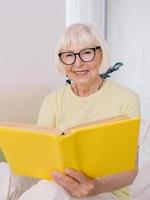 äldre kvinna med grått hår läser en bok på en soffa hemma. utbildning, pension, anti ålder, läsning koncept foto