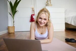 porträtt av skön blond kvinna ser på kondition Handledningar på bärbar dator, liggande på sudd yoga matta, följande träna instruktioner uppkopplad foto