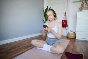 porträtt av ung blond kvinna i hörlurar, vändning på yoga, träna app på smartphone, välja musik på mobil telefon Ansökan, Sammanträde på sudd matta i rum foto