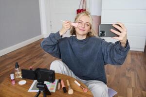 porträtt av skön ung livsstil bloggare, kvinna vlogger inspelning på digital kamera, leva ström smink handledning på smartphone app, Sammanträde i sovrum, använder sig av borsta på henne kinder foto