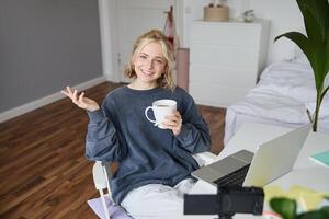 bild av ung kvinna, social media influencer, redigering henne på bärbar dator, sitter i en rum med dator och digital kamera, dricka kaffe, leende på kamera foto