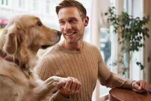 stänga upp porträtt av söt smart hund ger Tass till hans ägare. stilig man med hans gyllene retriever Sammanträde i en sällskapsdjur vänlig Kafé foto