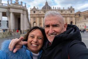 Lycklig mitten åldrig par på semester tar en selfie i främre av helgon peters basilika i rom foto