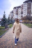 ung vuxen kvinna i jeans och sand täcka promenader genom de stad på en vår dag, modern arkitektur Bakom foto