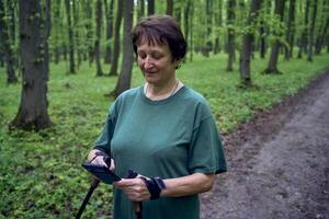 äldre kvinna håller på med nordic gående med pinnar i vår skog växlar podcast på telefon foto