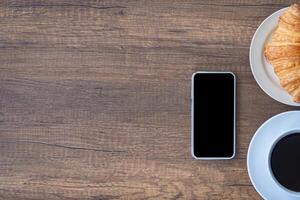 topp se av en skrivbord med en smartphone, en kopp av kaffe, och croissanter på en tallrik placerad på en trä- tabell. arbete från Hem. Plats för text foto