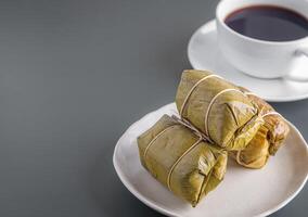 ångad klibbig ris med banan insvept banan blad på en tallrik med en vit kaffe kopp placerad på en grå bakgrund. traditionell thai mat stil. Plats för text foto