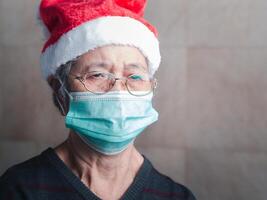 senior kvinna bär en kirurgisk mask på grund av till coronavirus och bär en santa claus hatt, ser på de kamera medan stående i de levande rum. Plats för text. begrepp av åldrig människor och jul foto