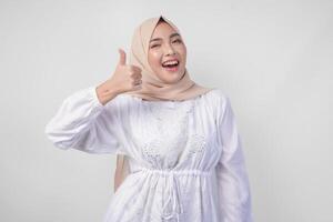 attraktiv ung asiatisk kvinna i vit klänning och hijab gestikulerar tummen upp för godkännande, Bra, excellent tecken, isolerat förbi vit bakgrund foto