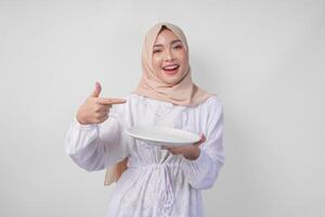leende ung asiatisk muslim kvinna i en slöja hijab pekande till ett tömma tallrik med kopia Plats över Det, presenter mat meny för iftar. ramadan begrepp foto