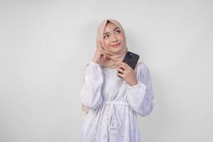 omtänksam ung asiatisk muslim kvinna bär vit klänning och hijab, använder sig av smartphone medan innehav henne haka och tänkande med allvarlig uttryck över isolerat vit bakgrund foto