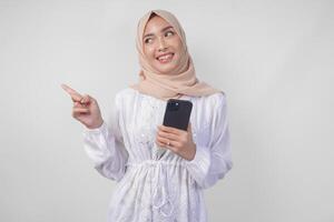 leende asiatisk kvinna bär hijab och vit klänning använder sig av smartphone medan pekande till de kopia Plats på henne rätt sida, stående över isolerat vit bakgrund. ramadan och eid mubarak begrepp foto