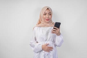 porträtt av ung asiatisk muslim kvinna i vit klänning och hijab känsla hungrig medan ser på henne telefon till beställa smaskigt mat efter fasta, isolerat på vit bakgrund foto