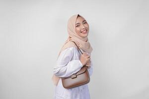 eleganta ung asiatisk muslim kvinna bär vit klänning och hijab med brun sling väska leende lyckligt över isolerat vit bakgrund. Ramadhan mode begrepp foto