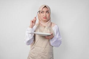 förvirrad ung asiatisk muslim kvinna i hijab och grädde förkläde innehav sked och tömma tallrik med kopia Plats över Det, tänkande hård Vad mat meny till äta för iftar. ramadan begrepp foto
