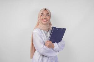 leende ung asiatisk muslim kvinna bär vit klänning och hijab innehav en dokumentera, isolerat förbi vit bakgrund. ramadan och eid mubarak begrepp foto