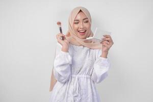 skön ung asiatisk muslim kvinna bär vit klänning och hijab applicering göra upp använder sig av borsta. mode och kosmetika skönhet begrepp foto