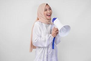 lycklig asiatisk kvinna bär hijab och vit klänning skrikande till de megafon framställning meddelande, stående över isolerat vit bakgrund foto