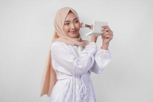 skön ung asiatisk muslim kvinna bär vit klänning och hijab applicering göra upp använder sig av borsta. mode och kosmetika skönhet begrepp foto