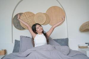 skön asiatisk kvinna vakna upp från sömn liggande på säng i sovrum, stretching händer och leende lyckligt foto