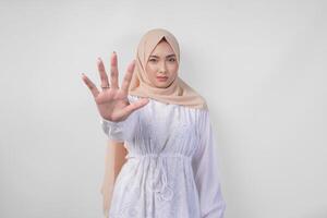 ung asiatisk muslim kvinna bär vit klänning och hijab som visar henne handflatan till göra en sluta gest med allvarlig uttryck foto