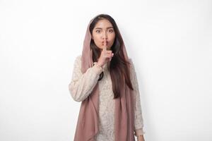 ung asiatisk muslim kvinna bär slöja slöja hijab ser till kamera medan sätta en finger i främre av mun gestikulerar stanna kvar tyst eller stanna kvar tyst, isolerat förbi vit bakgrund foto