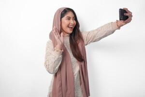 skön asiatisk muslim kvinna i slöja hijab slöja skrattande och tar en roligt selfie använder sig av mobil telefon, isolerat förbi vit bakgrund foto