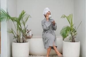 ung asiatisk kvinna dricka morgon- kaffe eller te i en kopp Sammanträde i en badkar bär grå morgonrock med isolerat grå bakgrund och krukväxter på både sidor foto