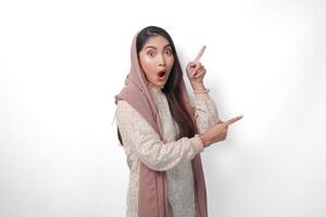 överraskad asiatisk muslim kvinna bär slöja slöja hijab ser chock medan pekande till kopia Plats bredvid henne, stående på isolerat vit bakgrund foto