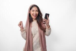 porträtt av ett upphetsad ung asiatisk muslim kvinna höjning näve upp gest, vinnande och fira seger utgör medan innehav en smartphone. ramadan och eid mubarak begrepp foto